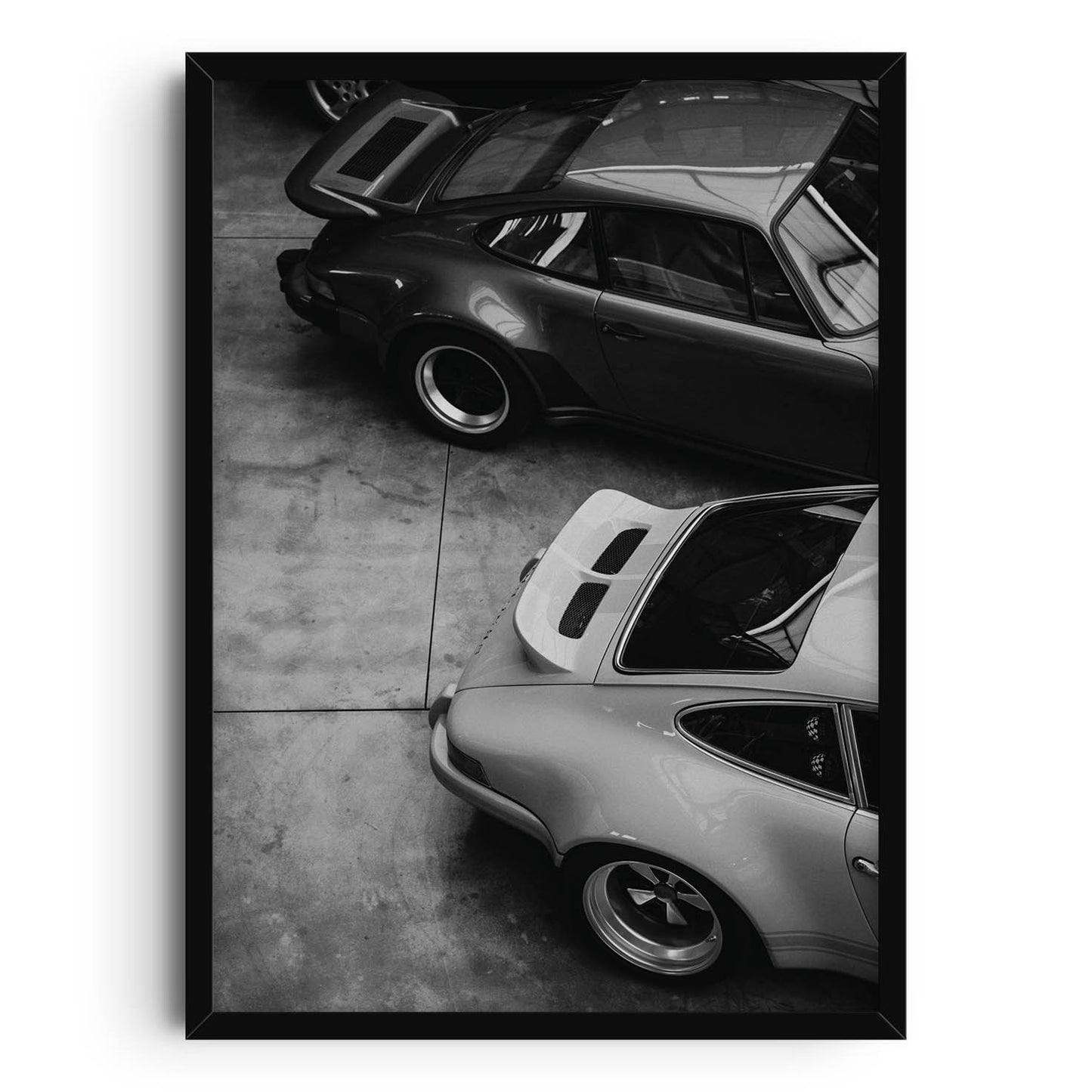 Print featuring a black-and-white photograph of two vintage sports cars parked in a garage. The top-down view showcases the sleek curves and distinct designs of the cars, highlighting their timeless, retro appeal. Perfect for modern home decor.