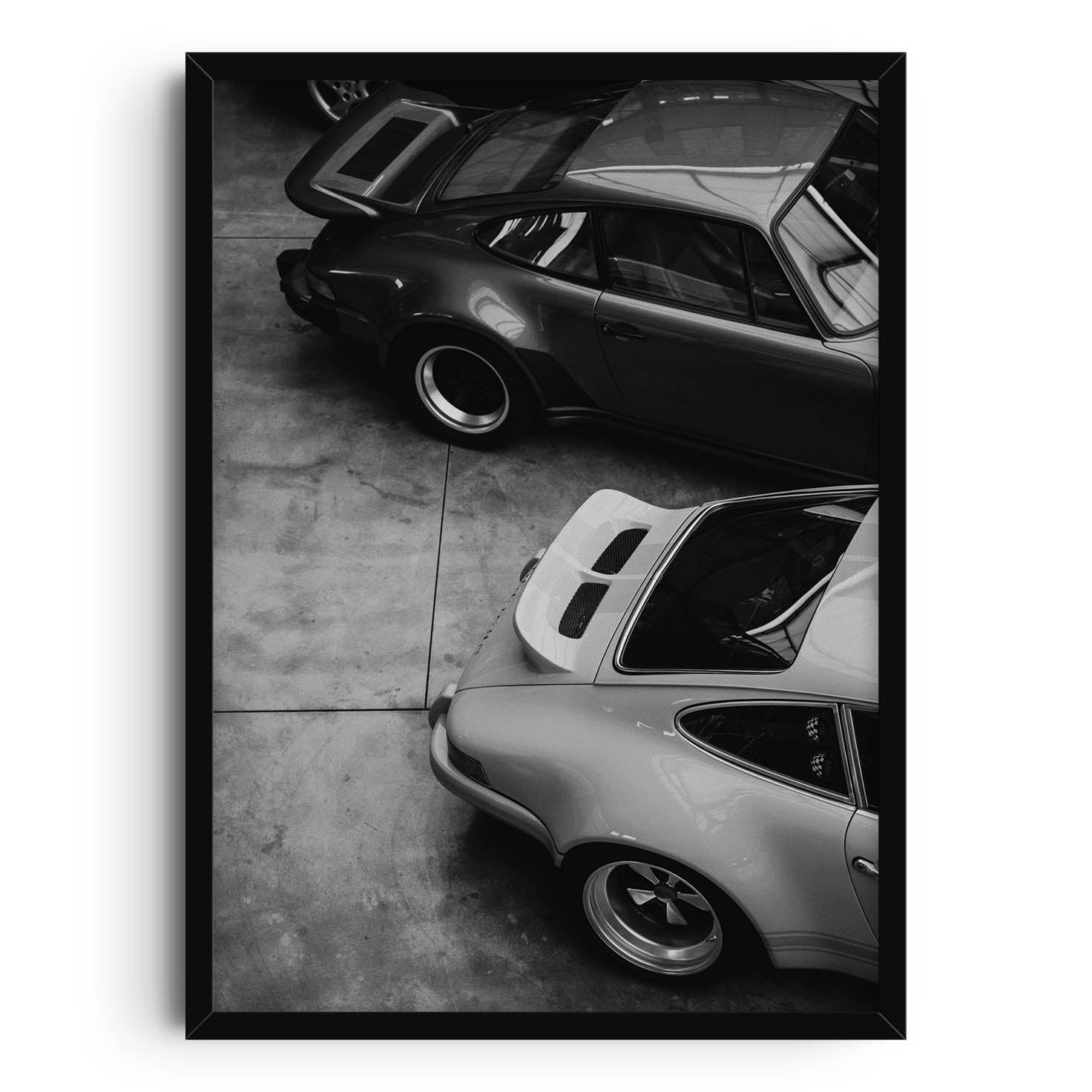 Print featuring a black-and-white photograph of two vintage sports cars parked in a garage. The top-down view showcases the sleek curves and distinct designs of the cars, highlighting their timeless, retro appeal. Perfect for modern home decor.
