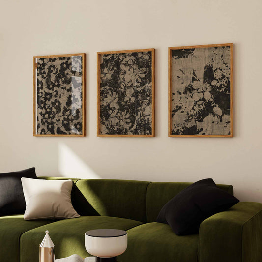 Three framed rustic floral pattern art prints in earthy tones hanging on a beige wall above a green velvet sofa with black and cream cushions, creating a cozy, nature-inspired interior setting.