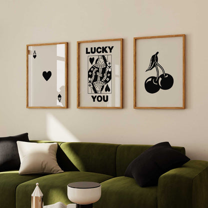 Set of three black and white art prints in wooden frames displayed on a beige wall above a green sofa. The artwork includes a playing card with an ace of spades, a 'Lucky You' card with a king of spades, and a minimalist cherry illustration.