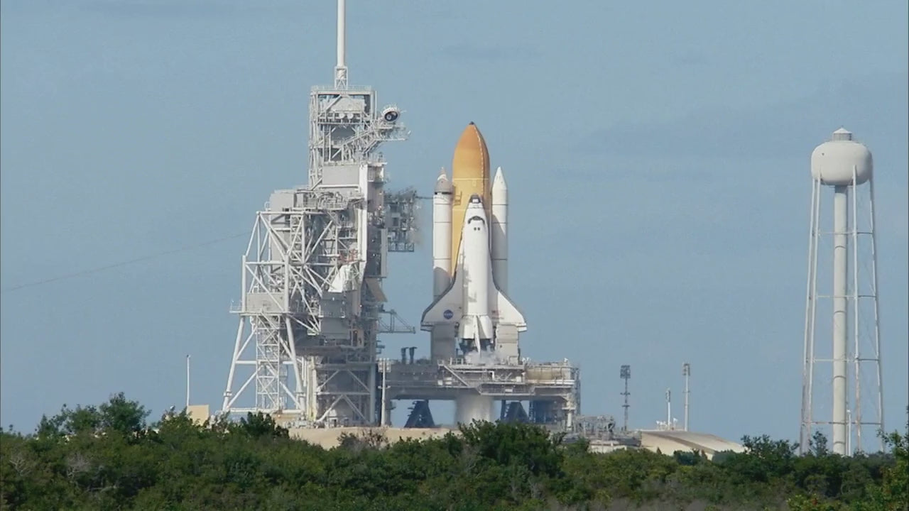 Video of a NASA Space Shuttle launching into the sky, showing fiery liftoff, plumes of smoke, onboard booster footage, and a wide shot of spectators watching near the countdown clock.